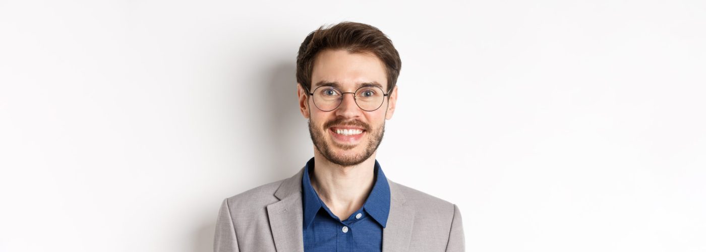 Close-up of handsome young office worker in glasses and suit smiling at camera, looking happy on white background.